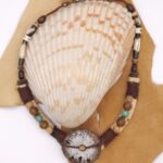 A hand carved sand dollar sits between different types of wooden beads as well as hairpipe and tiny shark vertebrae on a hemp cord and leather strap.