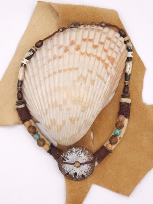 A hand carved sand dollar sits between different types of wooden beads as well as hairpipe and tiny shark vertebrae on a hemp cord and leather strap.