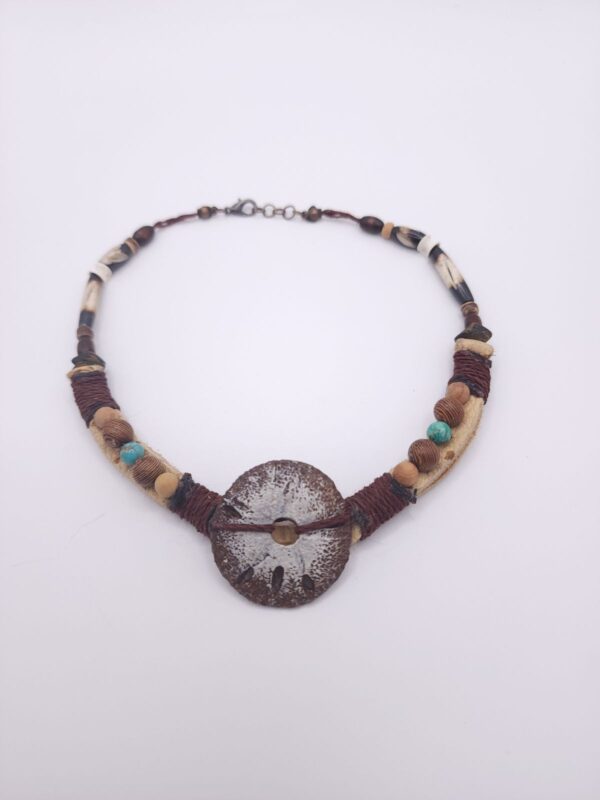 A hand carved sand dollar sits between different types of wooden beads as well as hairpipe and tiny shark vertebrae on a hemp cord and leather strap.