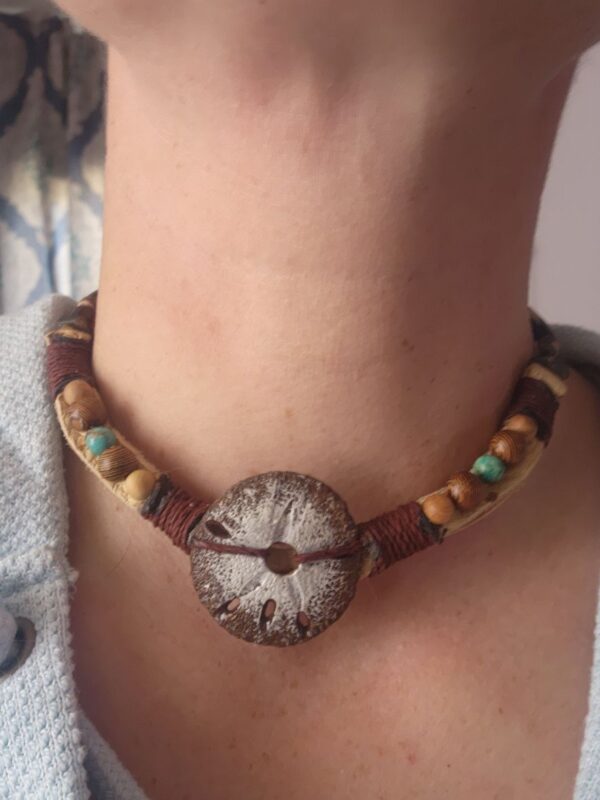 A hand carved sand dollar sits between different types of wooden beads as well as hairpipe and tiny shark vertebrae on a hemp cord and leather strap.