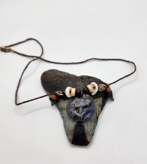 A shark carved into a coconut disc and painted gray as a pendant on a necklace with fossilized shark teeth, puka shells, and coconut beads