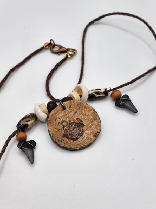 A shark carved into a coconut disc and painted gray as a pendant on a necklace with fossilized shark teeth, puka shells, and coconut beads