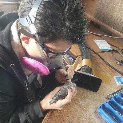 the artist is carving a small drinking horn while wearing a respirator at a desk using a dremel
